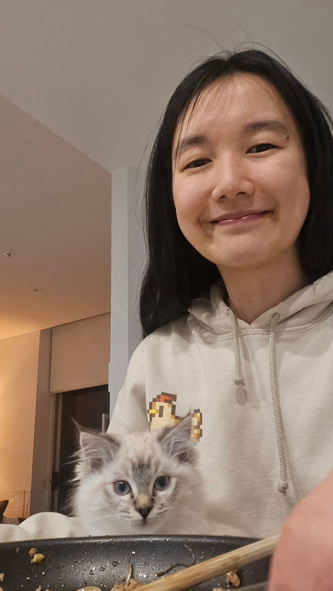 Selfie of Wing sitting at a kitchen island with a tabby ragdoll kitten on her lap and her food in a pan in the foreground. The kitten is looking straight at the camera with blue eyes. Wing is smiling while looking at the camera too.